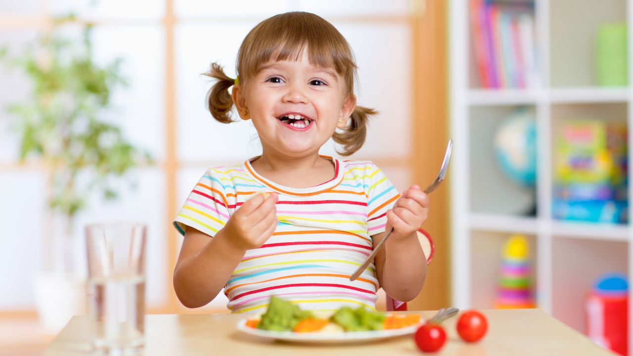 A smiling toddler holds a fork over a plate of vegetables at a small table.