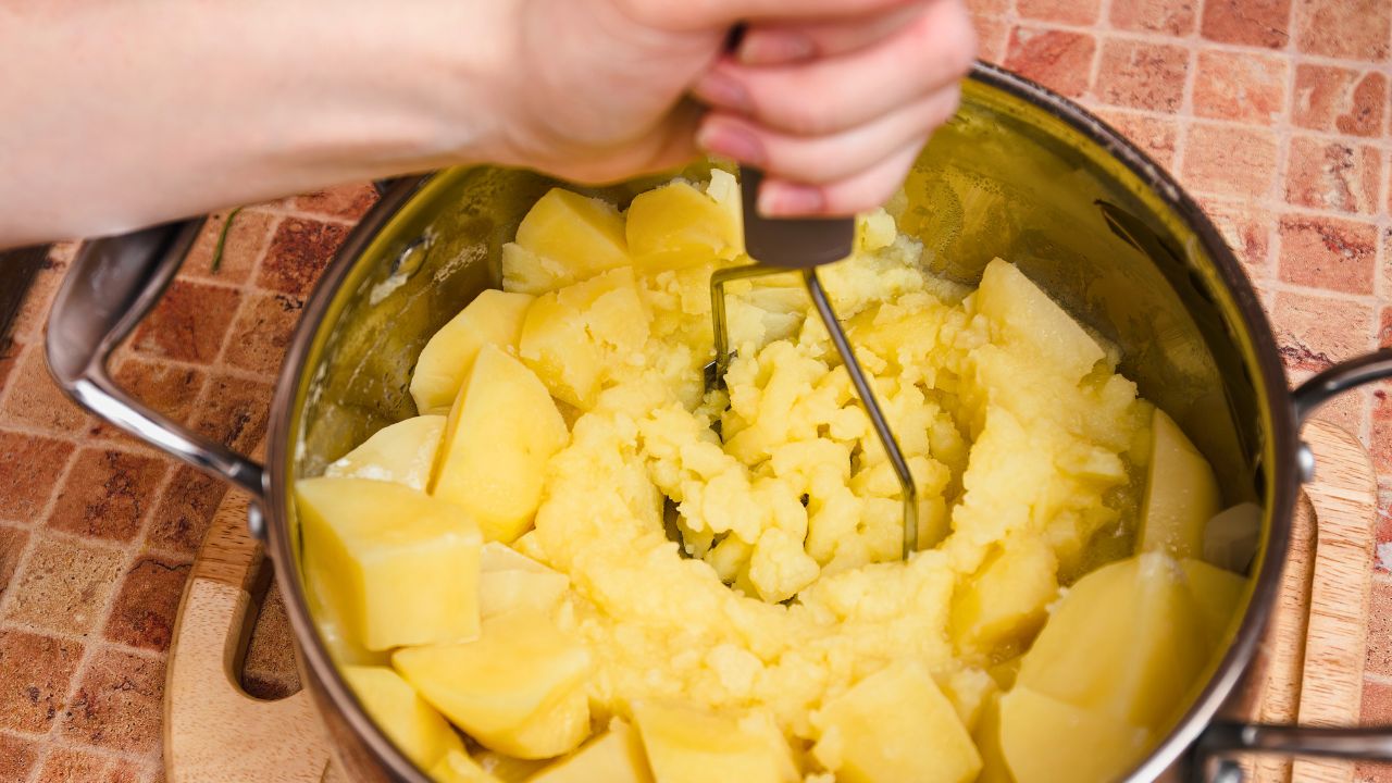 Hands mashing potatoes in a stainless-steel pot on a kitchen counter.