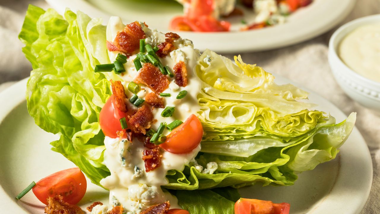 A wedge salad with lettuce, bacon, tomatoes, and creamy dressing on a white plate.