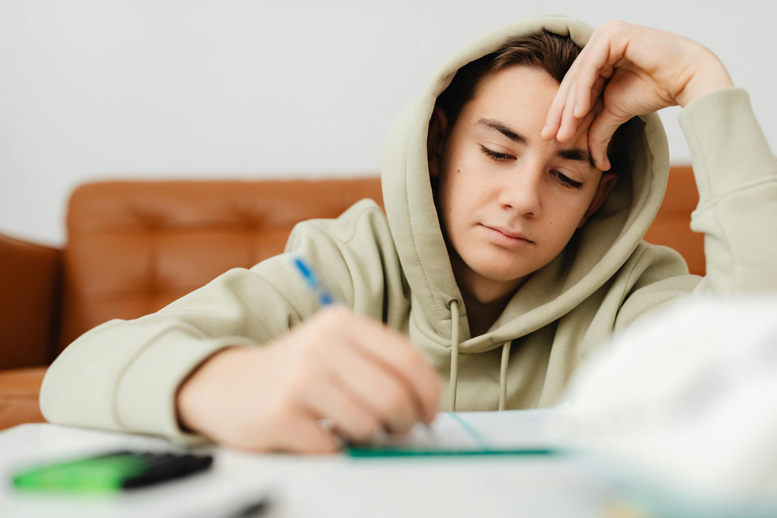 A young student wearing a hoodie intently studying at home, writing notes.