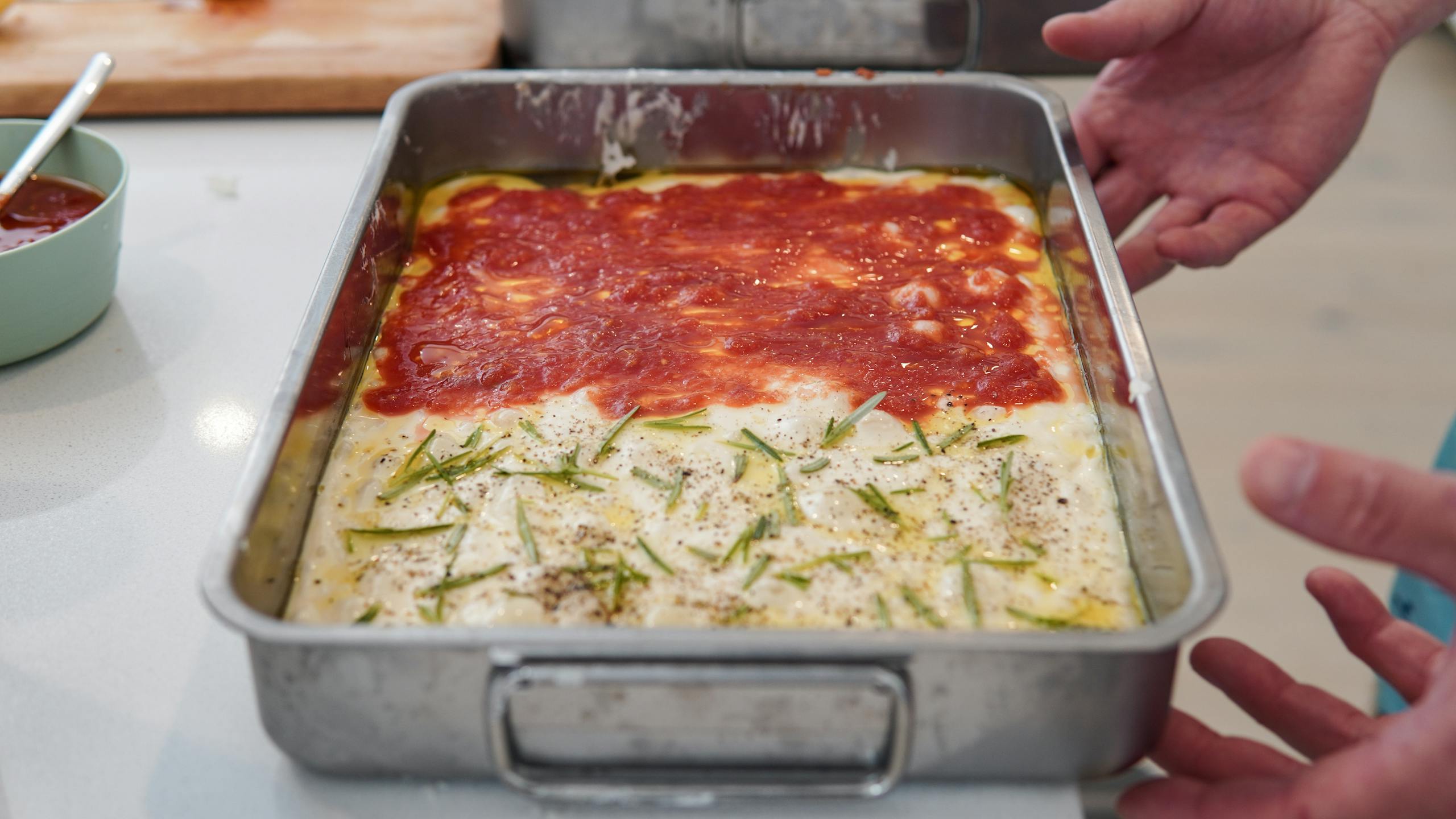 Hands holding a rectangular baking pan with a dish topped in red sauce and creamy herbs.