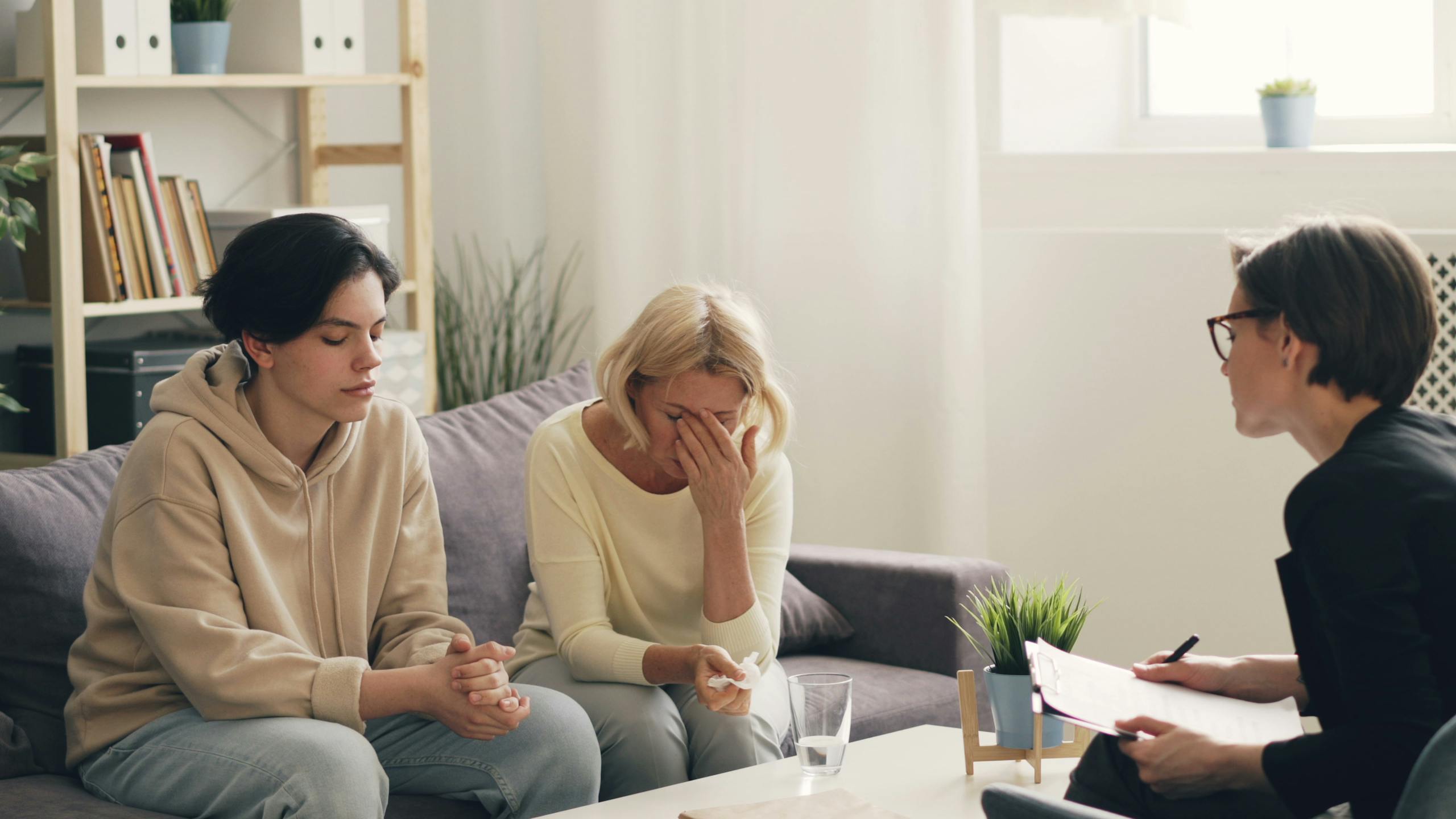 Three adults sit in a living room during a counseling session, one woman covering her face.