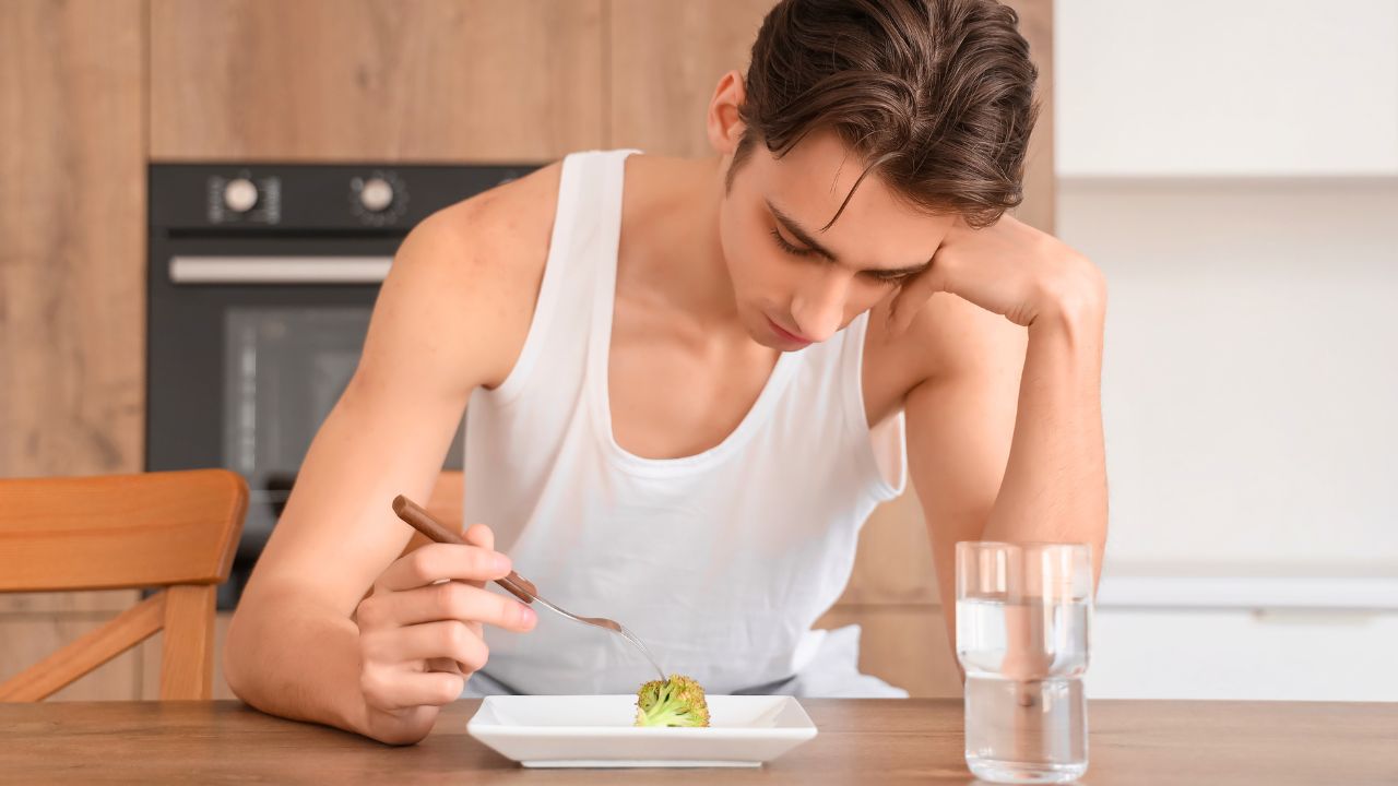 A young man in a white tank top sits at a table with a small piece of broccoli on a plate.
