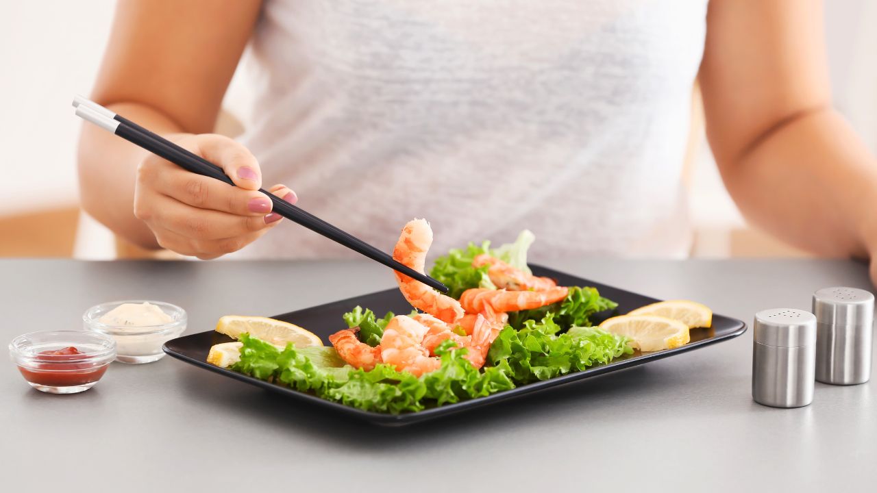 A person using chopsticks to pick up shrimp from a plate with lettuce and lemon wedges.