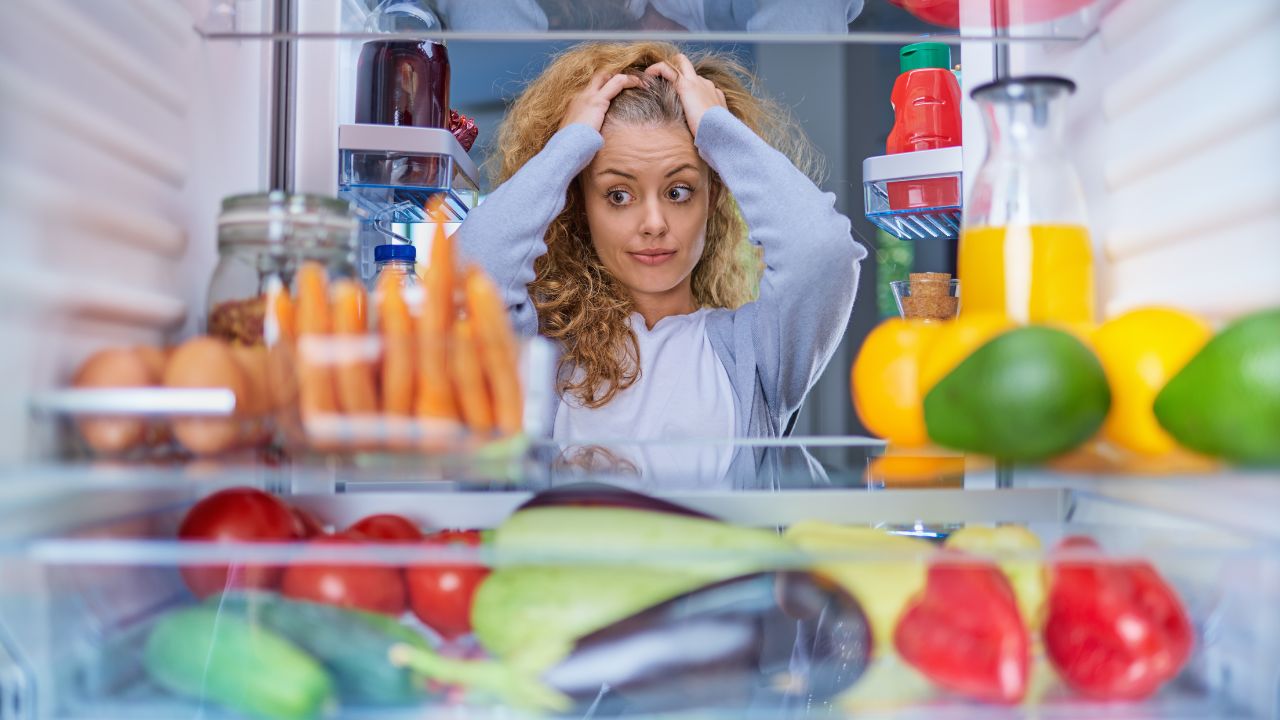 A woman stands in front of an open refrigerator looking stressed.