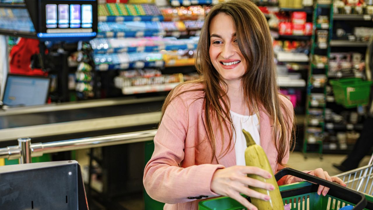 A smiling woman in a grocery store holds a banana over a shopping basket.