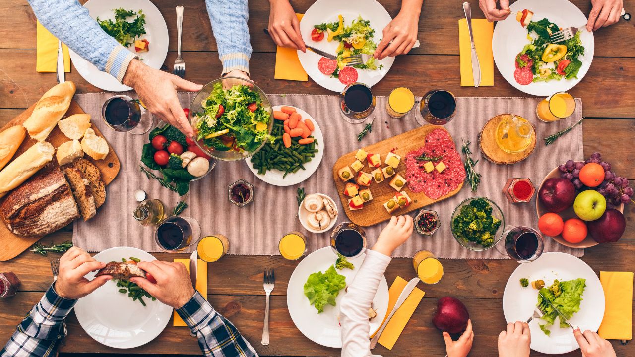 Overhead view of people sharing a meal with salads, bread, fruit, and drinks at a wooden table.