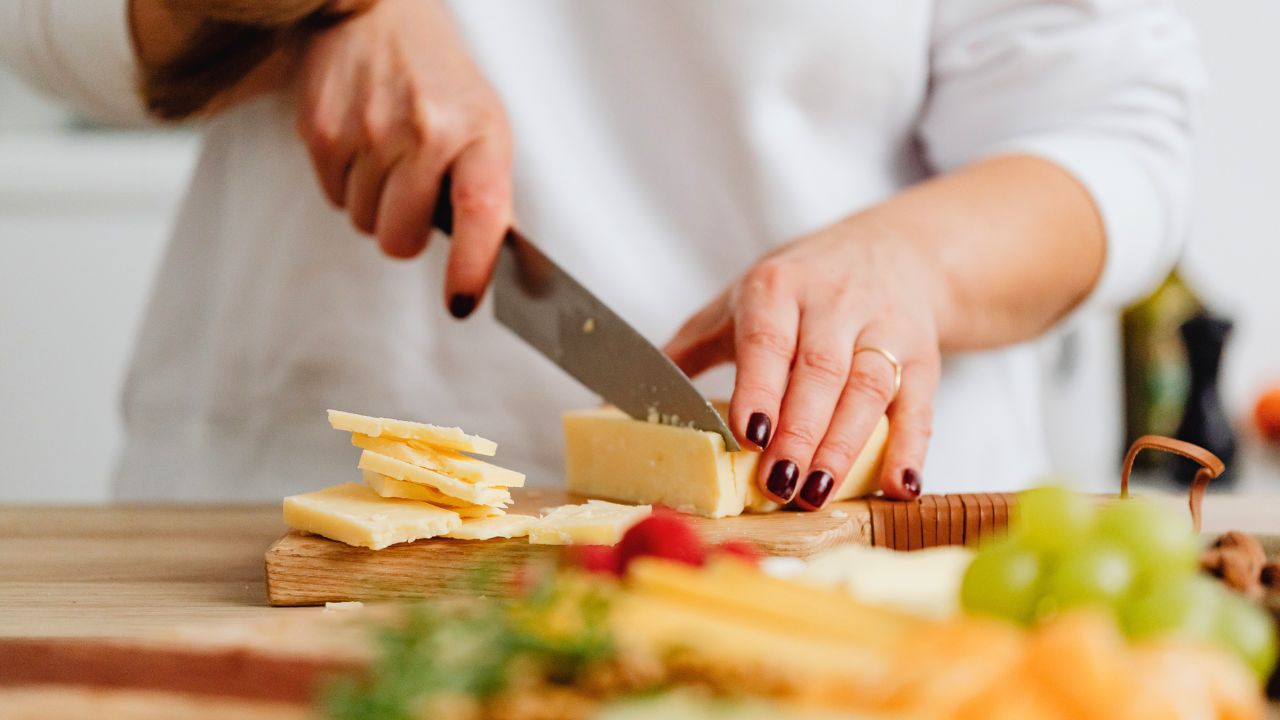 A person slices cheese on a wooden cutting board in a kitchen.