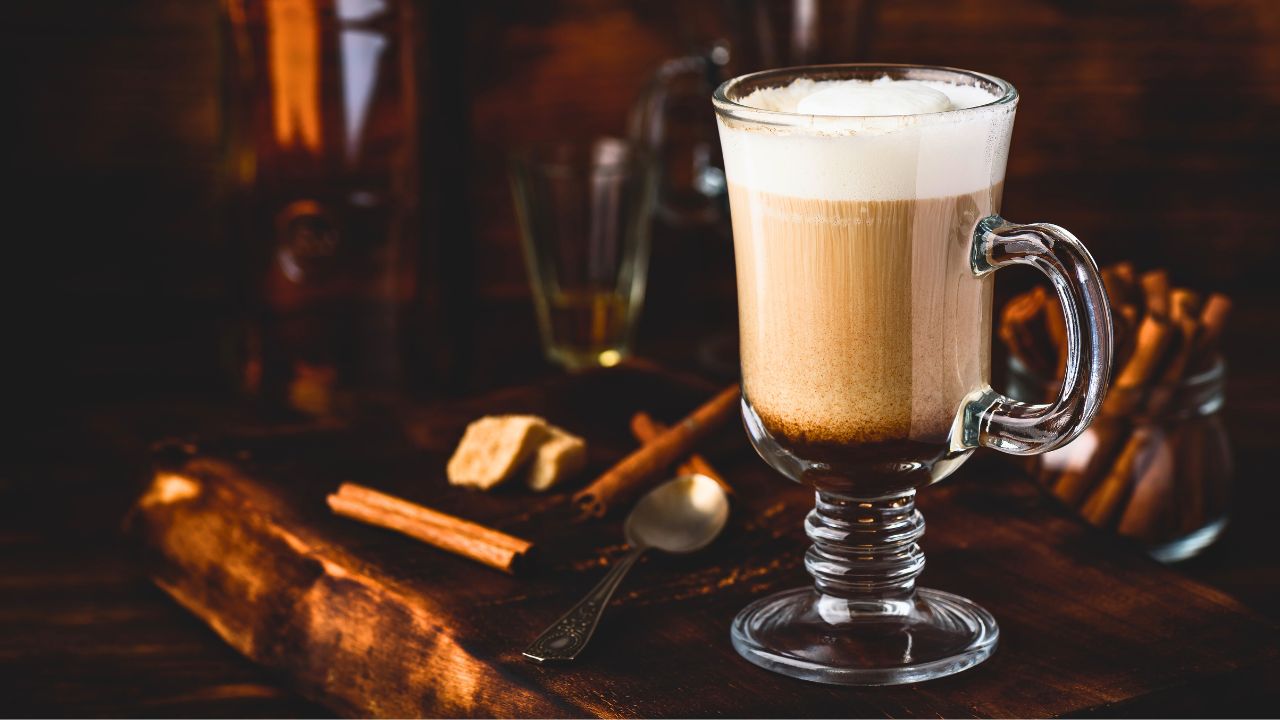 A frothy layered coffee drink in a glass mug on a wooden table with cinnamon sticks.