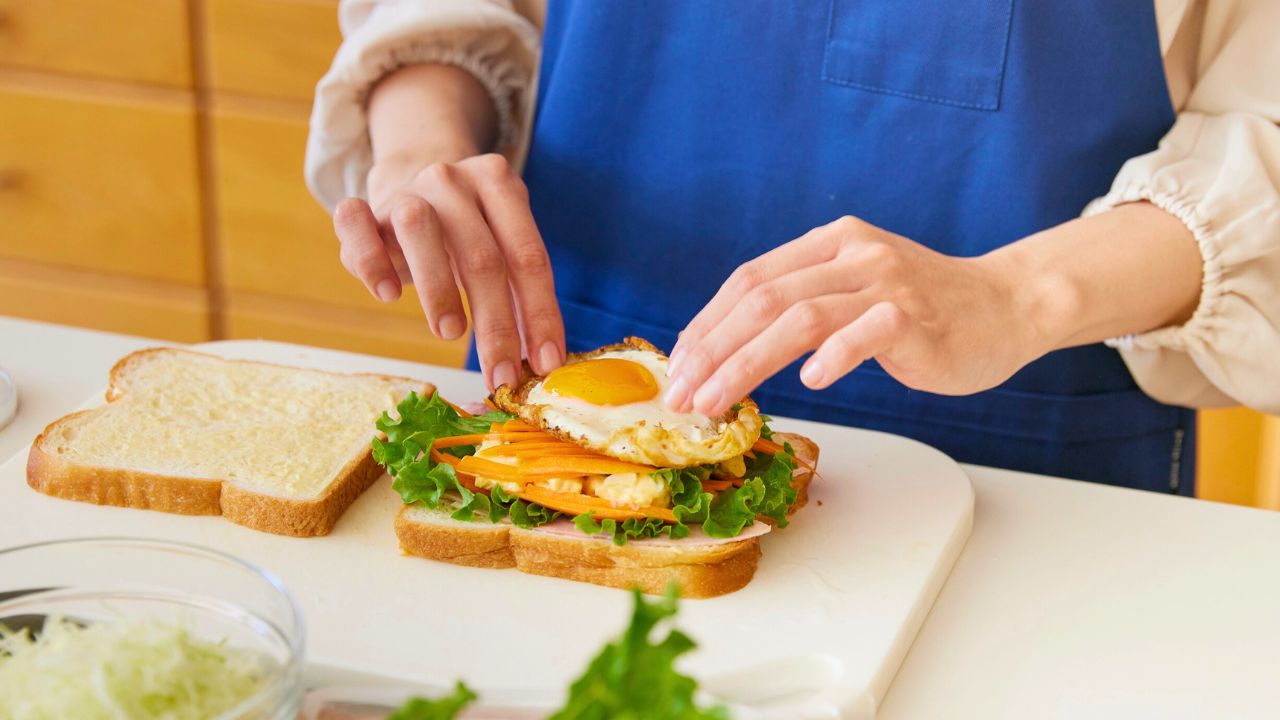 A person in a blue apron assembling a sandwich with lettuce, carrot, and a fried egg.