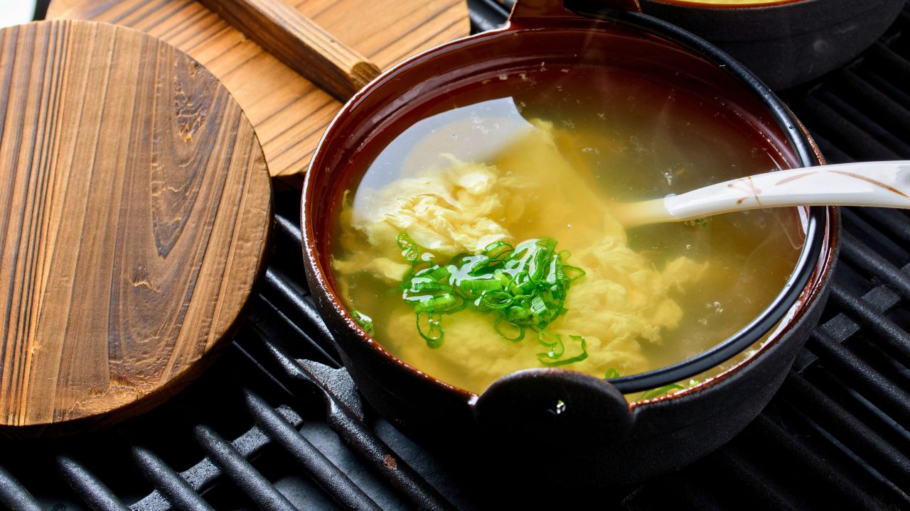 A bowl of egg drop soup with green onions and a spoon on a dark grill surface.