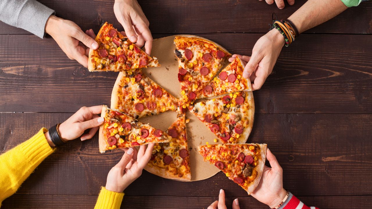 Several hands reach for slices of pizza on a wooden table.