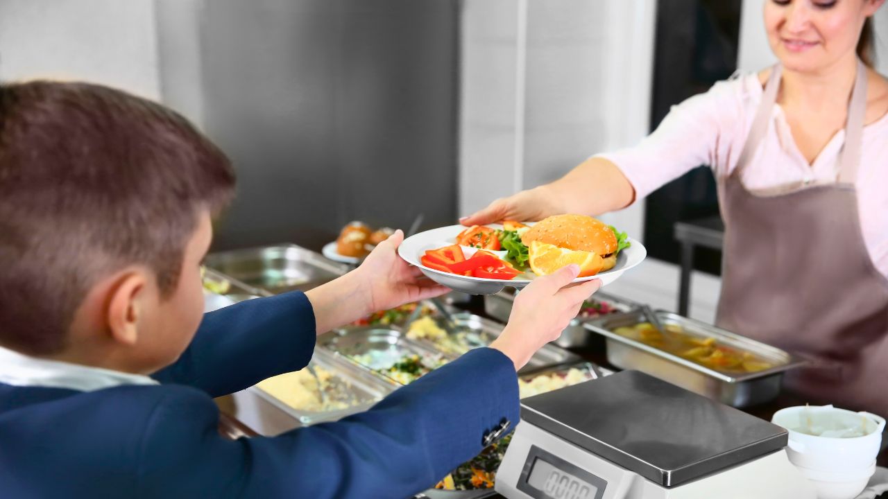 A boy receives a plate with a burger and vegetables from a woman at a buffet counter.