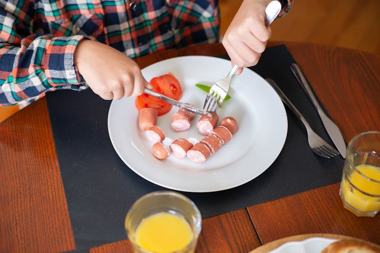 A child slices sausages at a breakfast table with juice, showing a cozy morning meal scene.