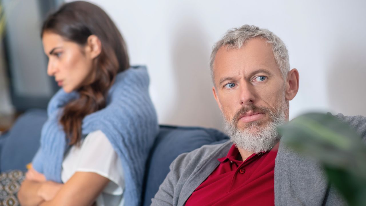 A tense couple sits apart on a couch in a living room.