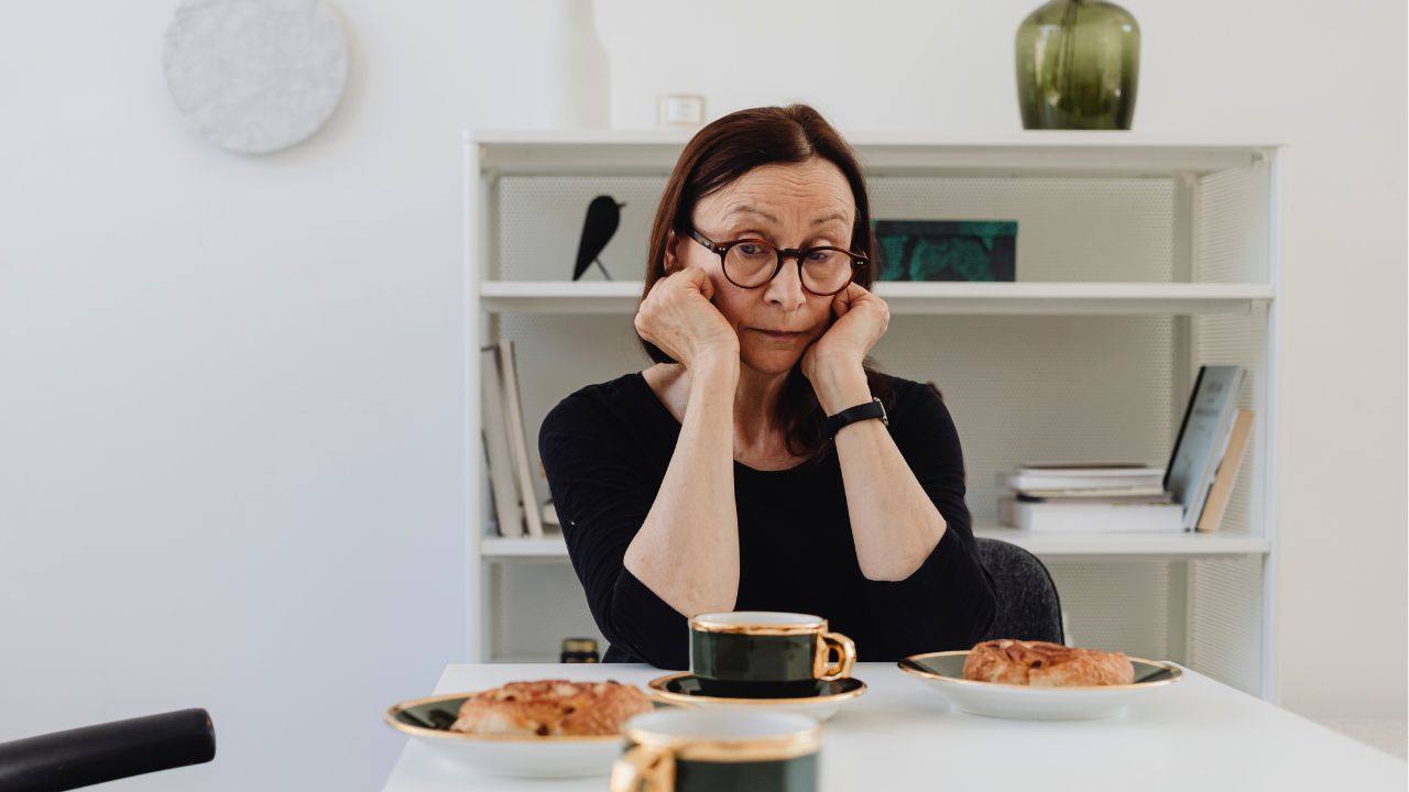A woman sits at a table with coffee and pastries, resting her face in her hands.
