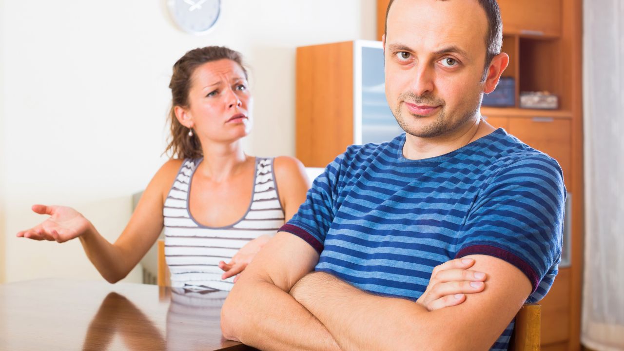 A man with crossed arms sits at a table while a woman gestures behind him in a kitchen.
