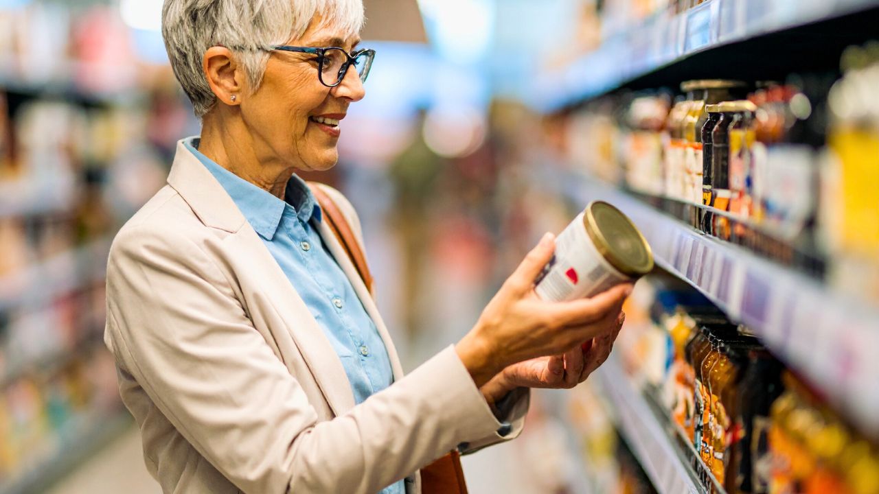 An older woman smiles while holding a can in a supermarket aisle.