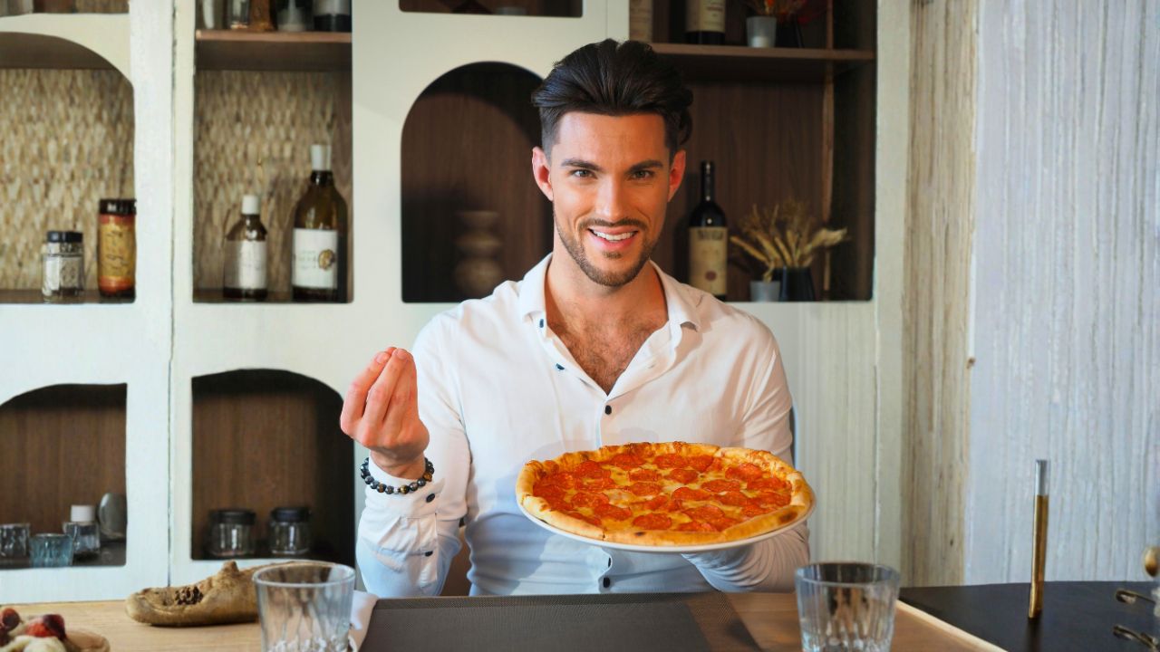 A smiling man holds a pepperoni pizza while seated at a dining table.
