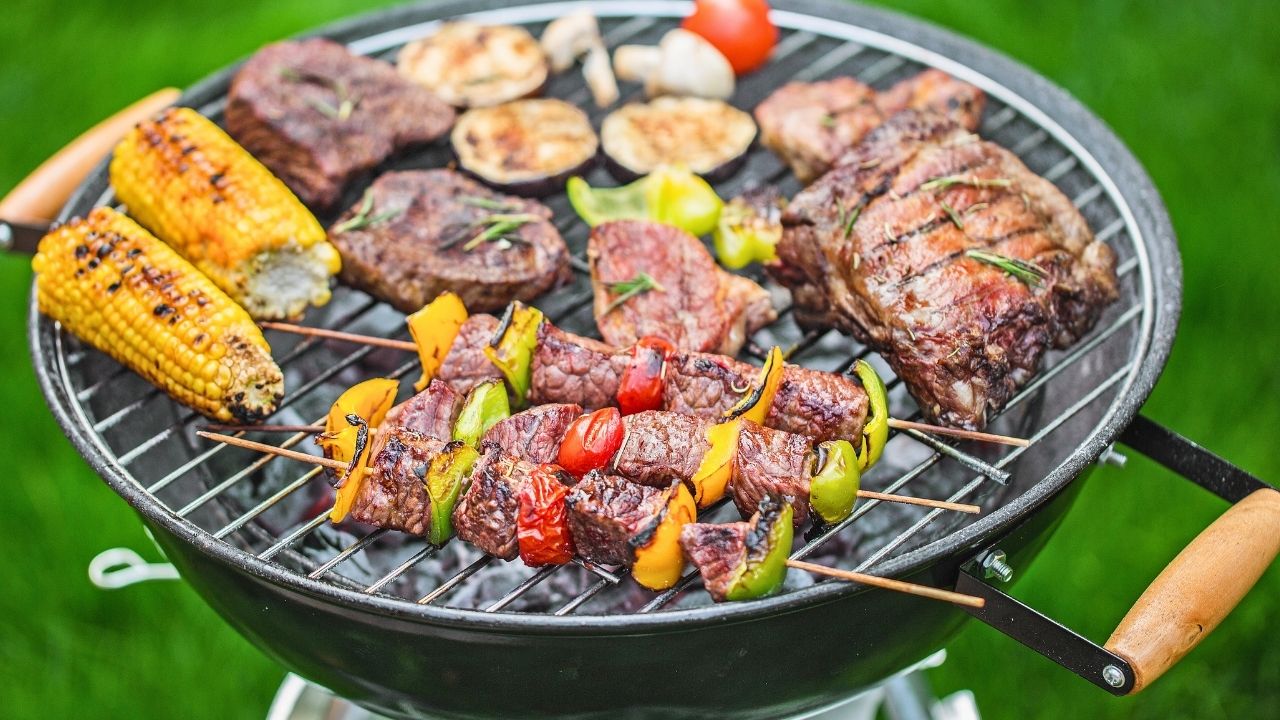 A backyard charcoal grill filled with steaks, vegetable skewers, and corn on the cob.
