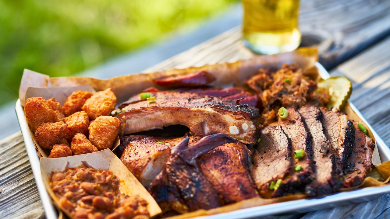 A tray of barbecue meats, fried bites, and baked beans on a picnic table.