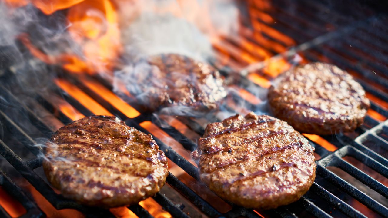 Four hamburger patties sizzling on a grill over open flames.