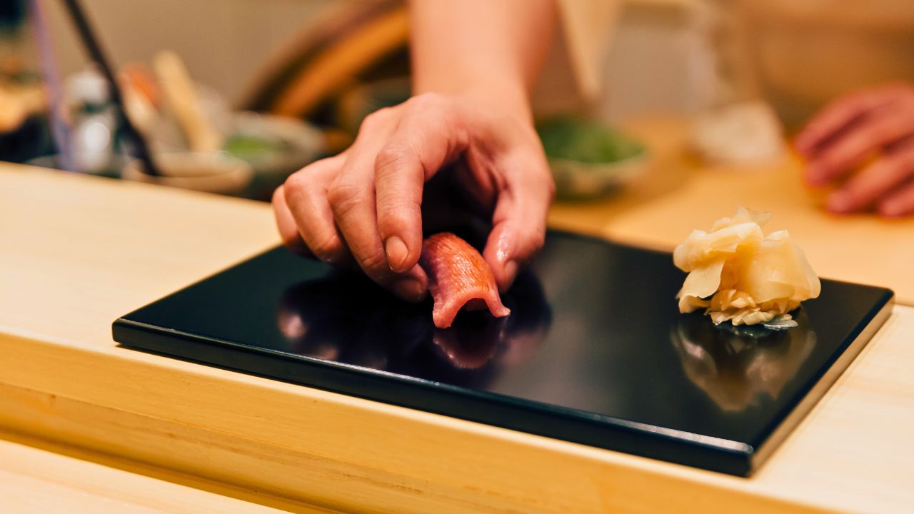A hand places a piece of sushi on a black plate beside pickled ginger.