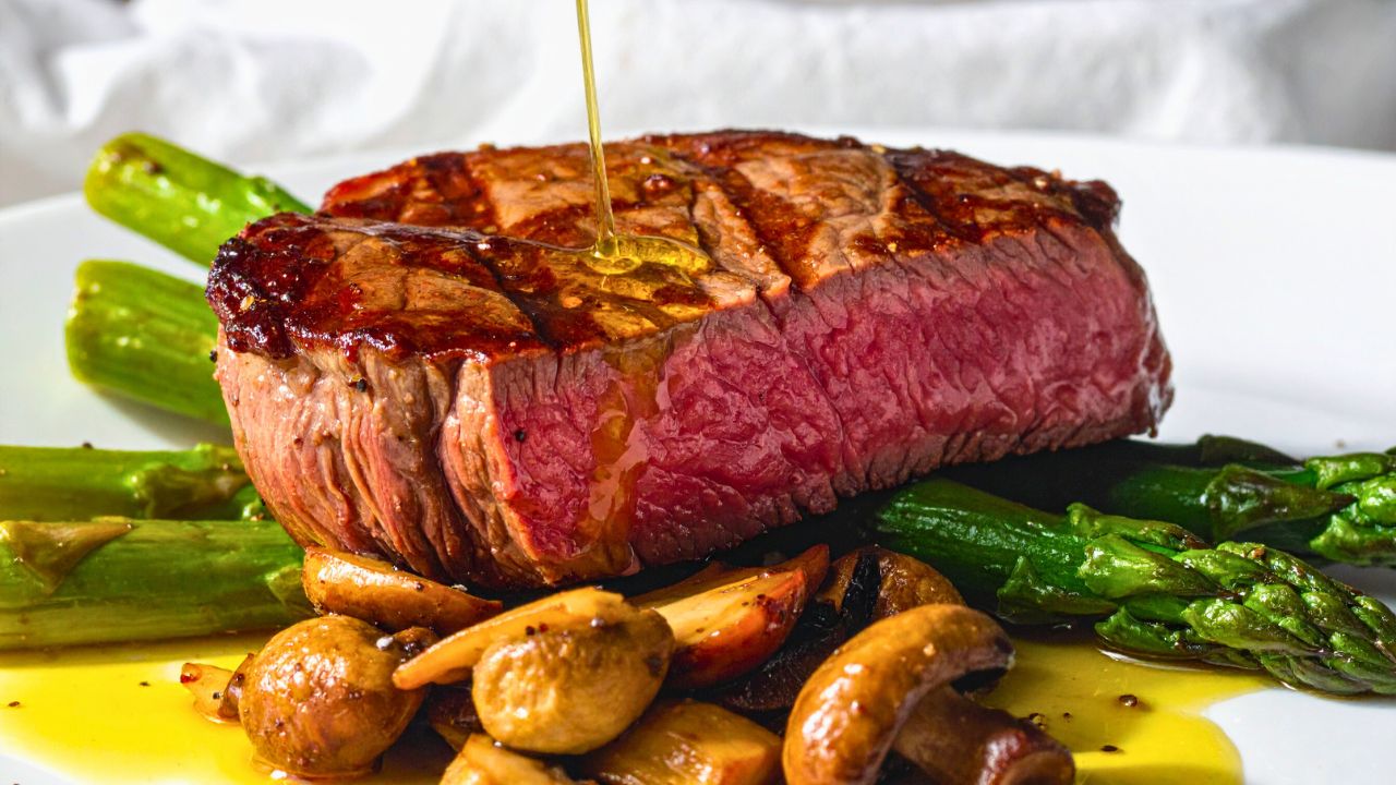 A medium-rare steak with asparagus and mushrooms on a white plate being drizzled with oil.
