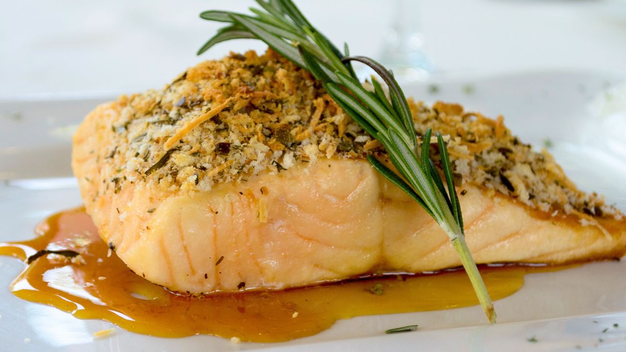 A breaded salmon fillet garnished with rosemary on a white plate with sauce.