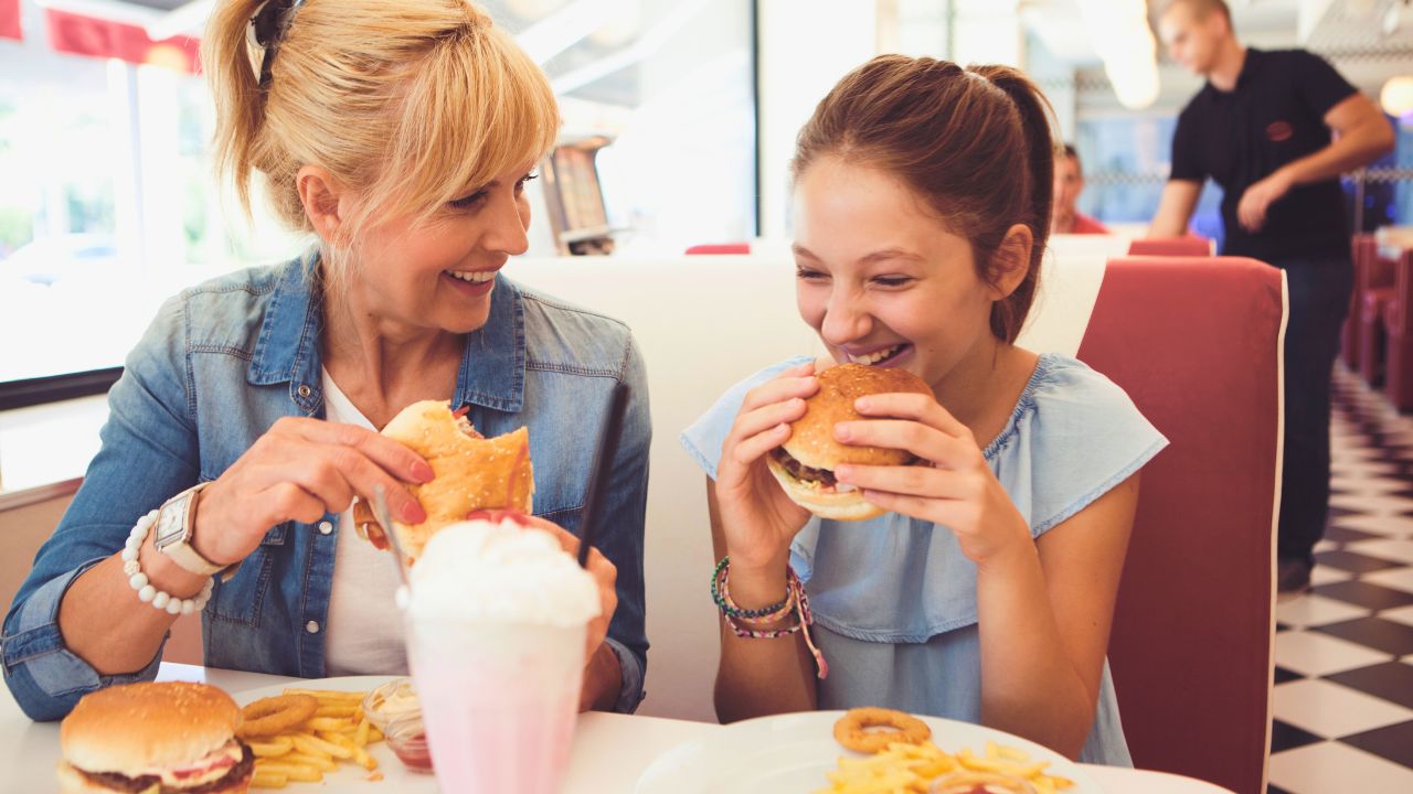 A mother and daughter smiling while eating burgers at a diner booth.