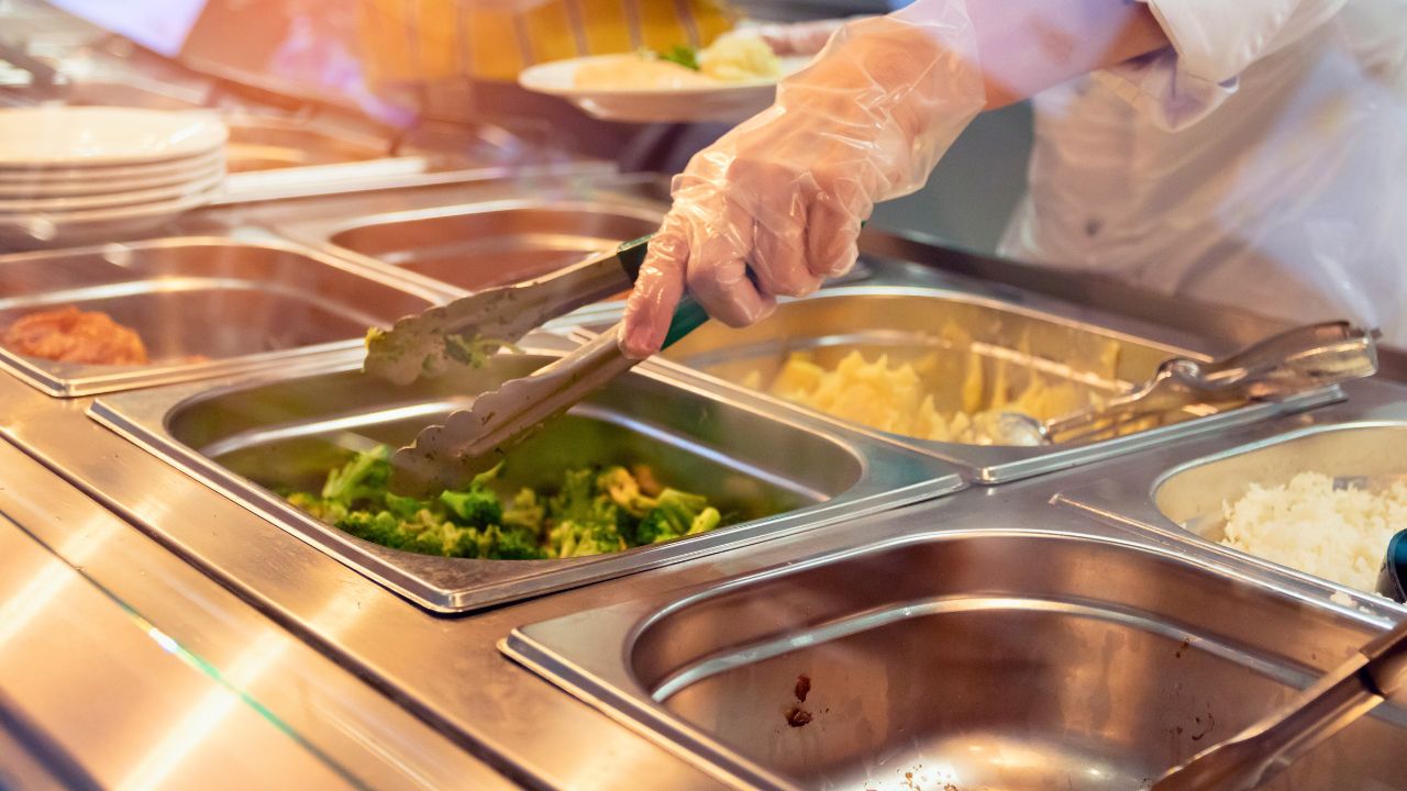 A gloved worker serves broccoli from a stainless-steel buffet tray.