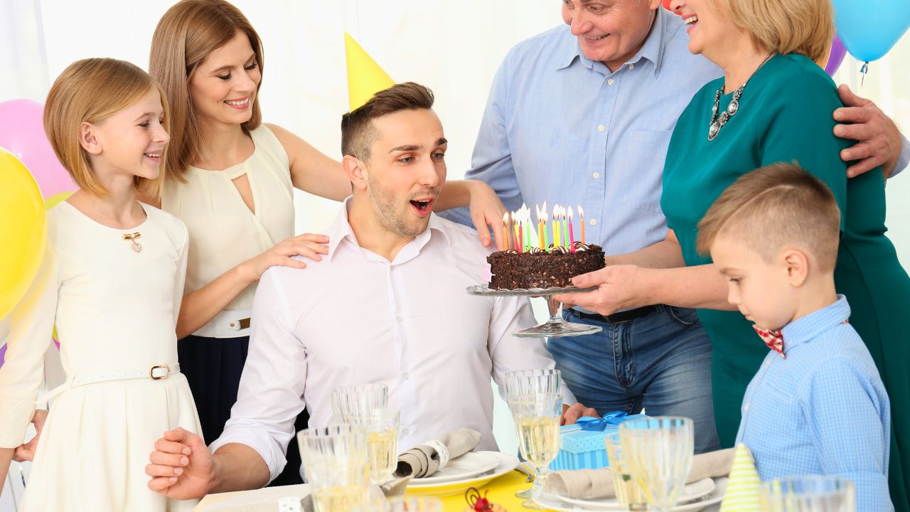 A family presents a lit chocolate birthday cake to a seated young man at a party table.