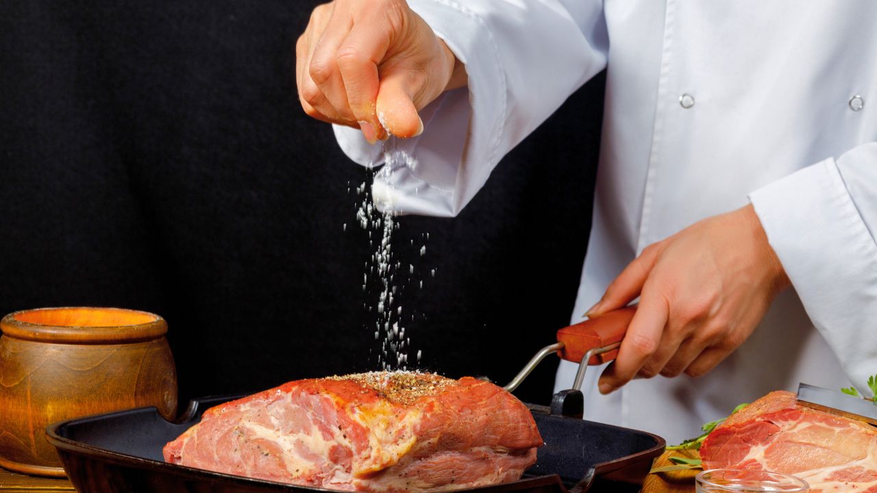 A chef sprinkles salt over raw meat in a black pan.