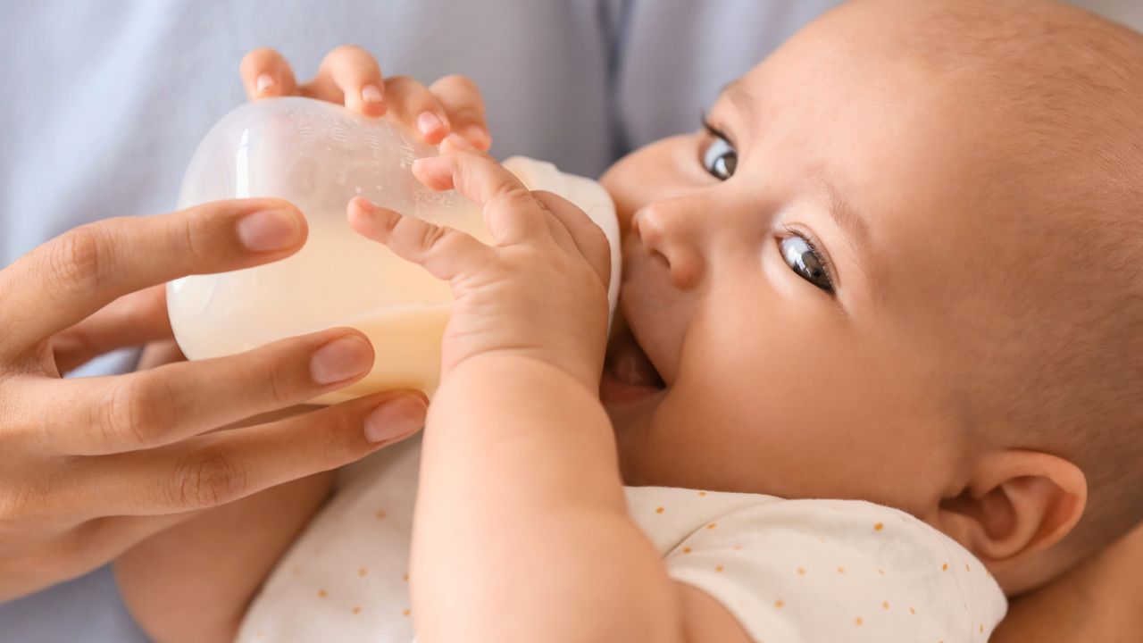 An adult hand holds a baby bottle while a baby drinks from it.