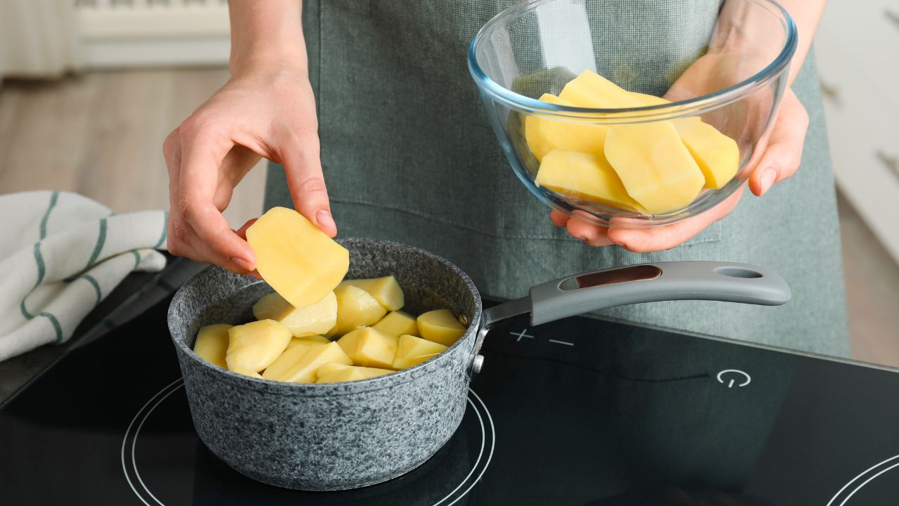 A person adds peeled potato pieces from a glass bowl into a saucepan on an induction cooktop.