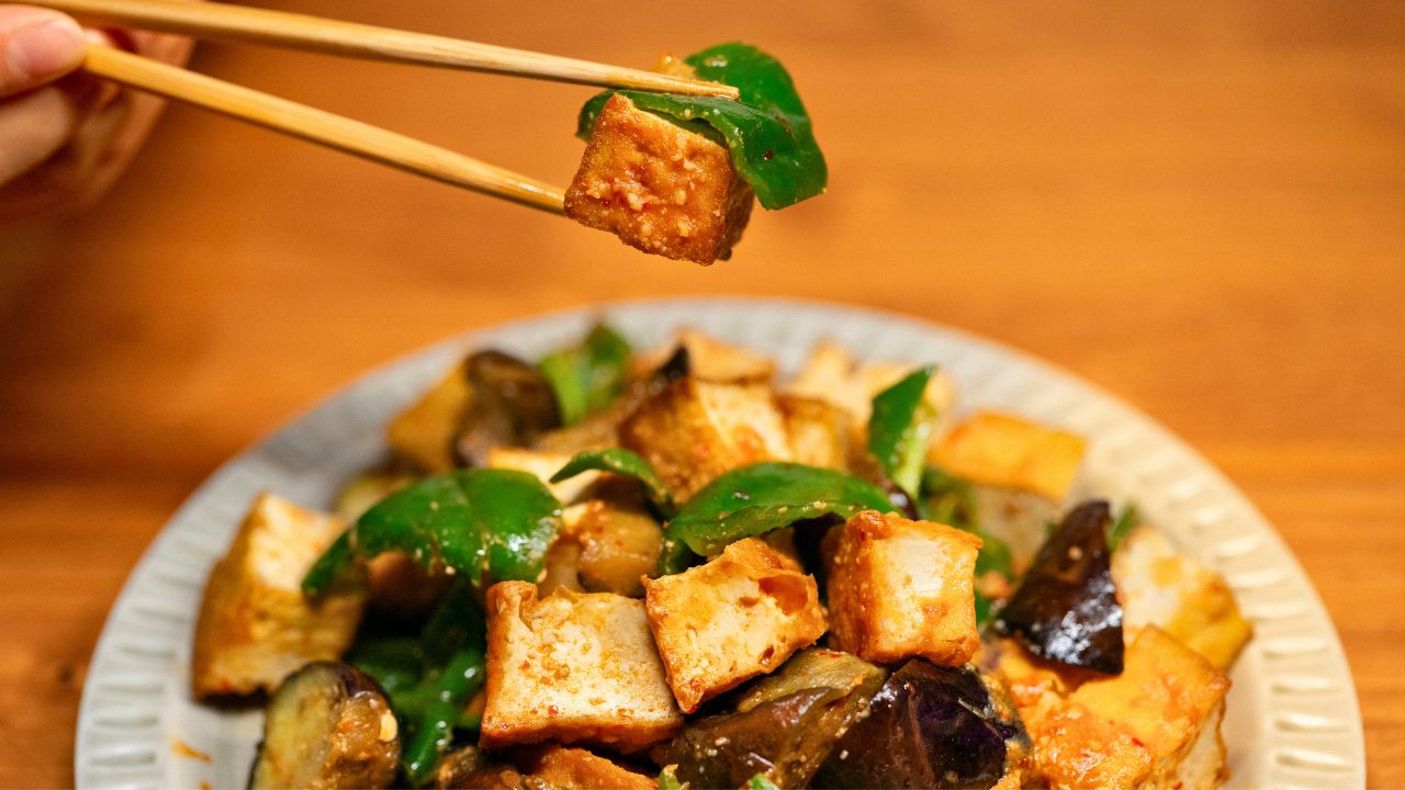 Chopsticks lifting a piece of tofu above a plate of stir-fried vegetables.